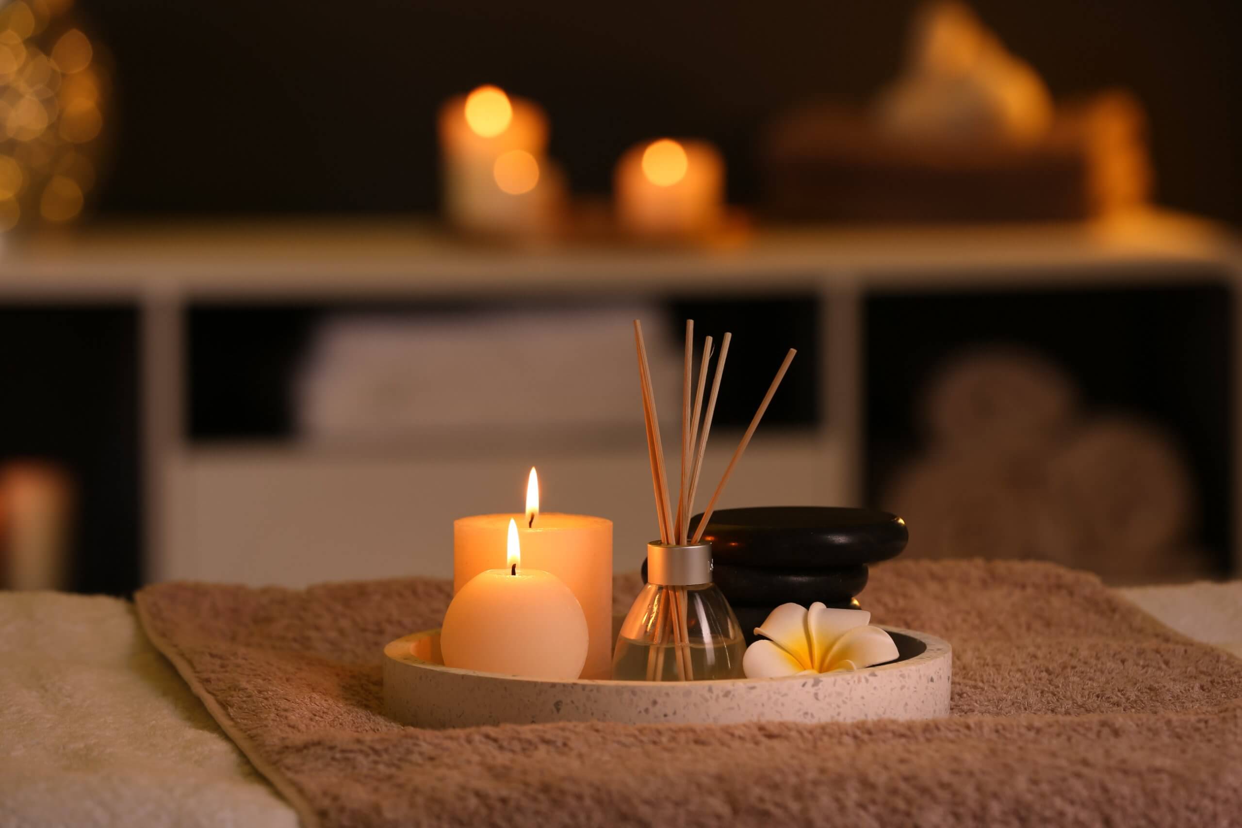 Ein rundes Tablett mit einer brennenden Kerze, einem Schilfdiffusor, schwarzen Steinen und einer Plumeria-Blüte auf einem Handtuch, mit verschwommenen Kerzen im Hintergrund.