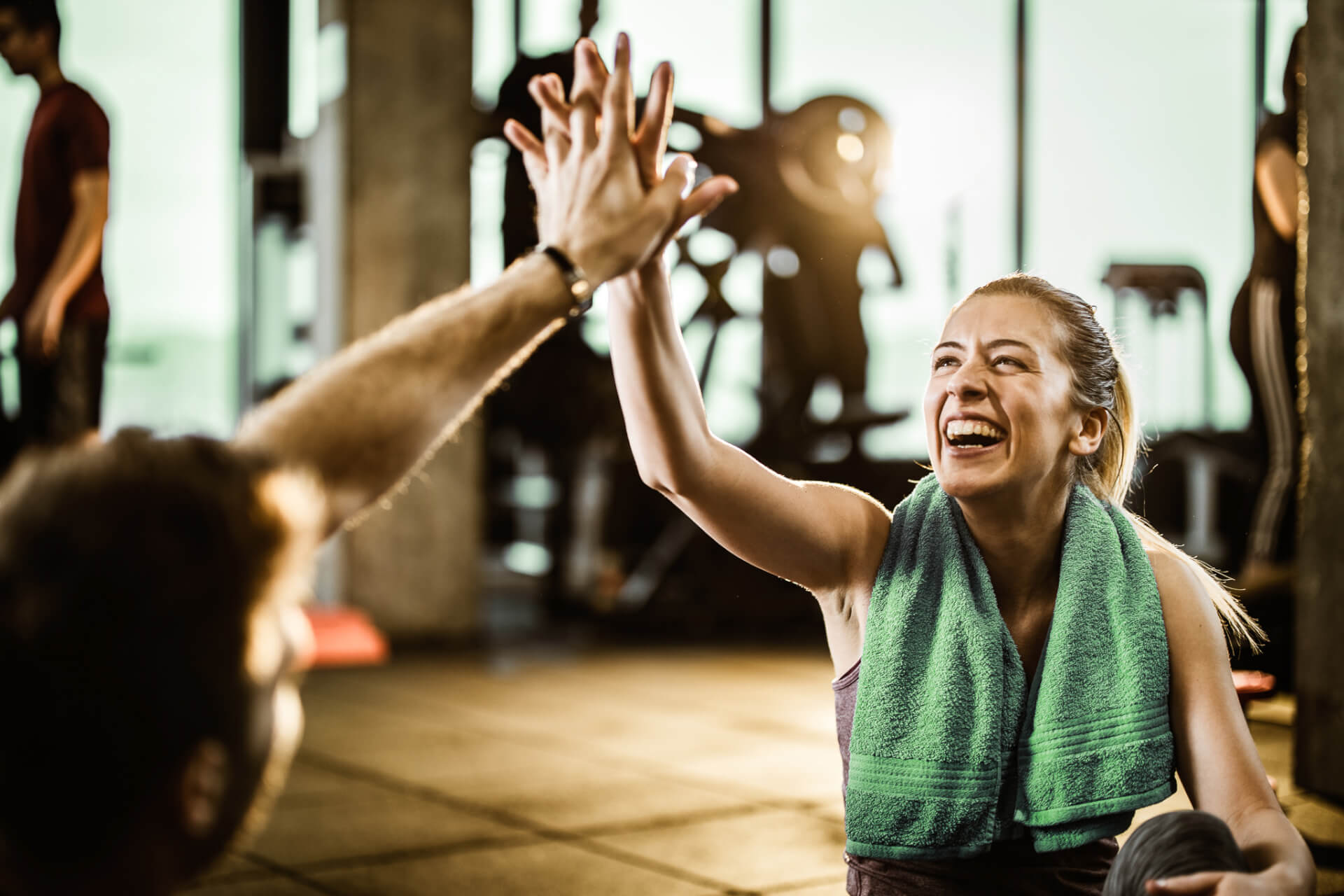 Une femme avec une serviette autour du cou sourit et fait un signe de la main à une autre personne dans un gymnase.