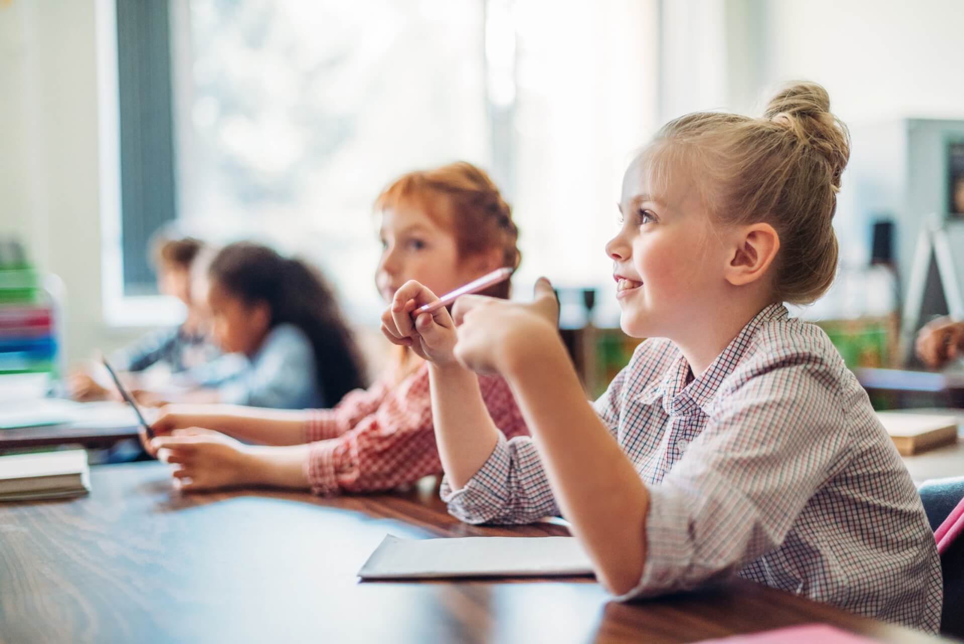 Les enfants sont assis à des pupitres dans une salle de classe, écoutant attentivement et tenant des crayons, avec des cahiers devant eux.
