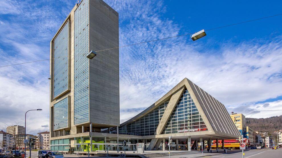 Un bâtiment moderne avec une haute tour rectangulaire et une structure de verre triangulaire adjacente sur fond de ciel bleu avec des nuages.