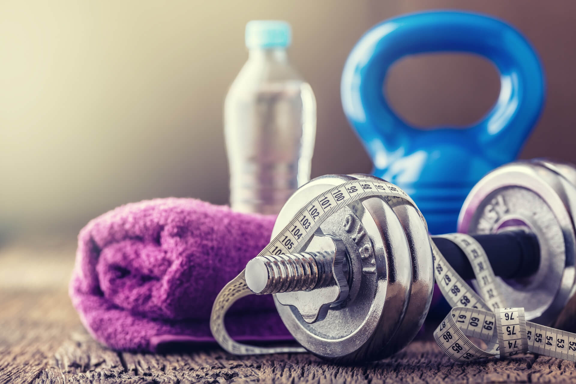 Eine Hantel, ein Maßband, ein lilafarbenes Handtuch, eine Wasserflasche und eine blaue Kettlebell sind auf einer Holzfläche angeordnet.