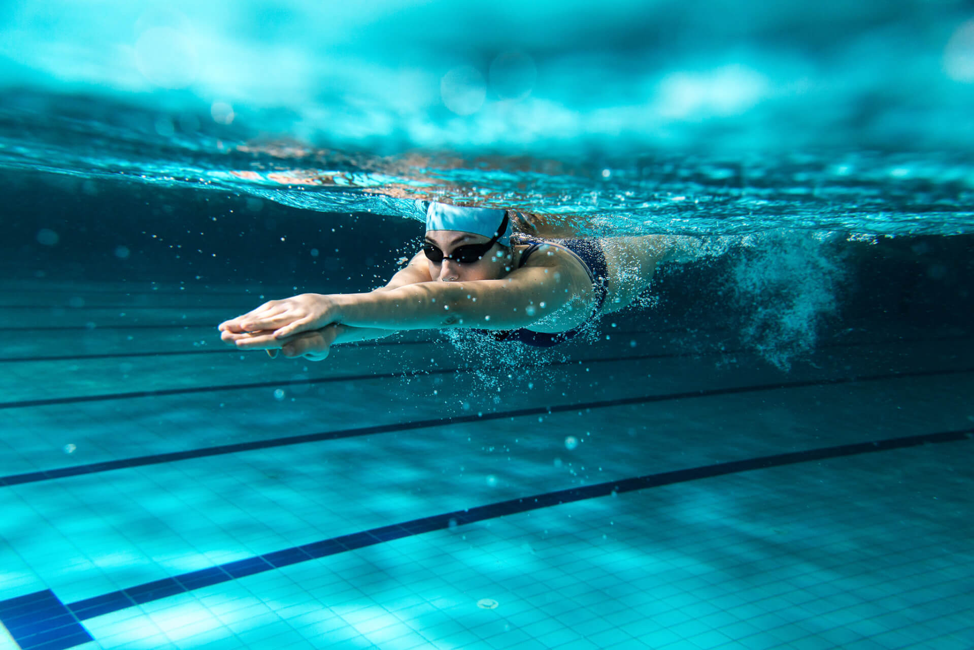 Vue sous-marine d'un nageur portant des lunettes et un bonnet de bain, glissant vers l'avant dans une piscine dont les couloirs sont visibles sur le sol carrelé.