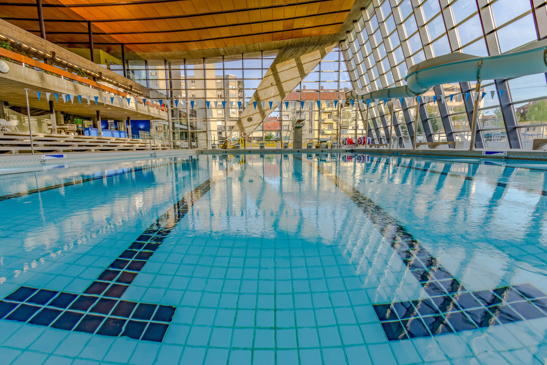 Piscine couverte à l'eau bleue claire, avec marquage des couloirs et toboggan aquatique, entourée de grandes baies vitrées et d'un plafond en bois.