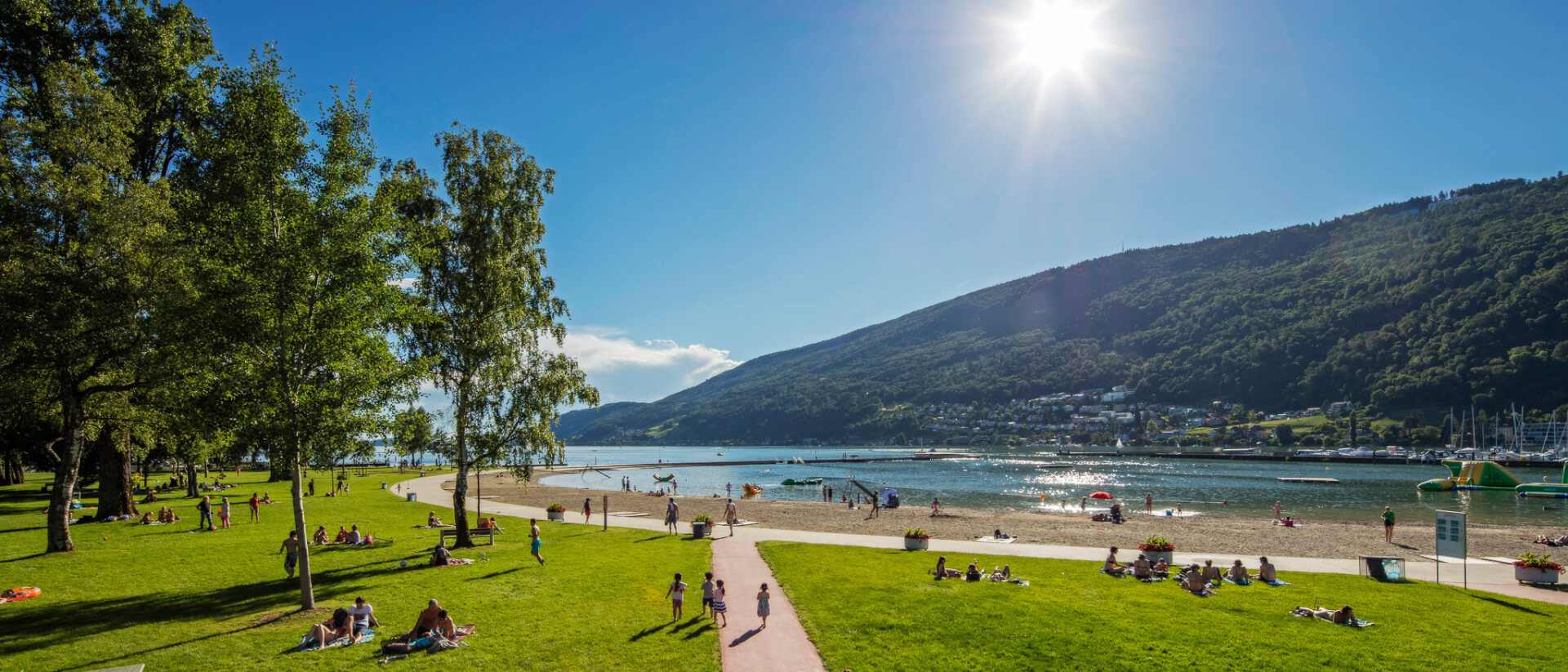 Menschen entspannen sich in einem grasbewachsenen Park an einem See, mit Sandstrand, Bäumen und fernen Hügeln unter einem strahlenden Sonnenhimmel.