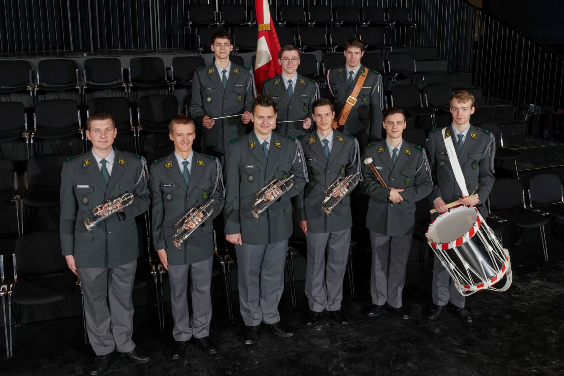 Neuf jeunes hommes en uniforme se tiennent à l'intérieur, tenant des cuivres, un tambour et un drapeau, disposés en deux rangées avec des sièges en gradins vides derrière eux.