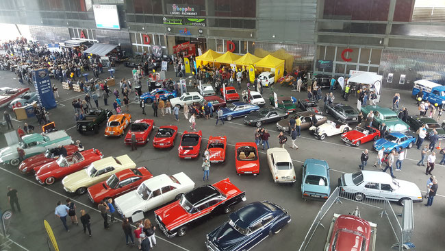 Vue aérienne d'un salon de l'automobile classique à l'intérieur, avec diverses voitures de collection exposées et des gens qui se promènent autour des stands des vendeurs et des expositions.