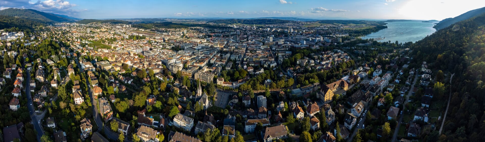 Vue aérienne d'un paysage urbain avec des bâtiments groupés, des espaces verts et un lac sur la droite, entouré de collines et de montagnes lointaines sous un ciel partiellement nuageux.