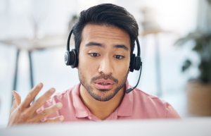 Un homme portant un casque et une chemise rose parle tout en faisant un geste de la main lors d'un appel vidéo.