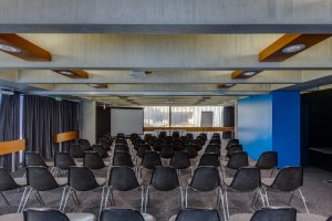 In einem Konferenz- oder Vortragsraum sind Reihen leerer schwarzer Stühle vor einem Projektionsschirm und einem langen Tisch an der Stirnseite angeordnet. Der Raum hat Betondecken und eine moderne Einrichtung.