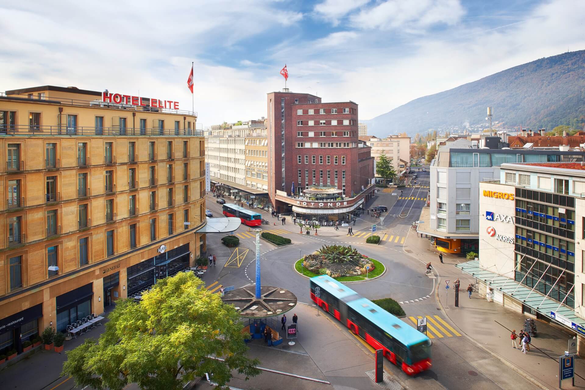 Un rond-point de ville avec des bus rouges, entouré de bâtiments comprenant l'hôtel Elite, des magasins et des piétons, avec une colline en arrière-plan sous un ciel partiellement nuageux.