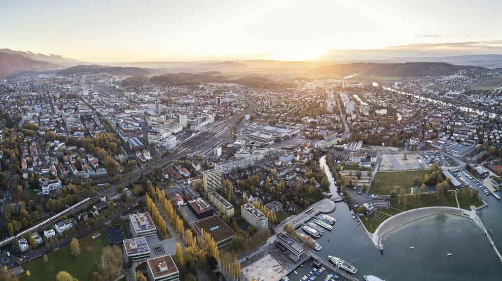 Vue aérienne d'une ville au coucher du soleil, montrant un mélange de bâtiments, de rues, un port de plaisance avec des bateaux, et les collines environnantes en arrière-plan.