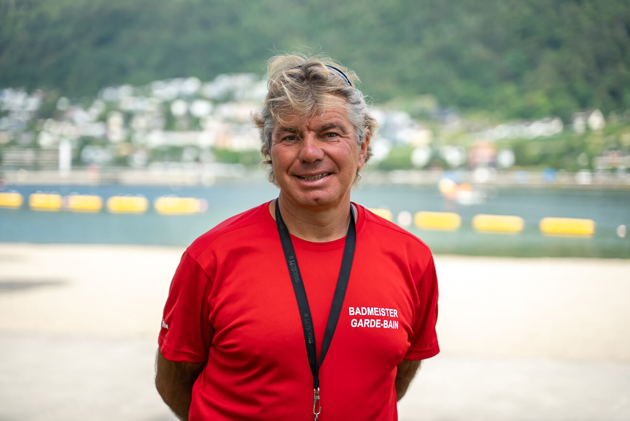 Ein Mann mit einem roten Badmeister Garde-Bain-Hemd und einer Pfeife steht im Freien am Wasser, mit Bergen und Gebäuden im Hintergrund.