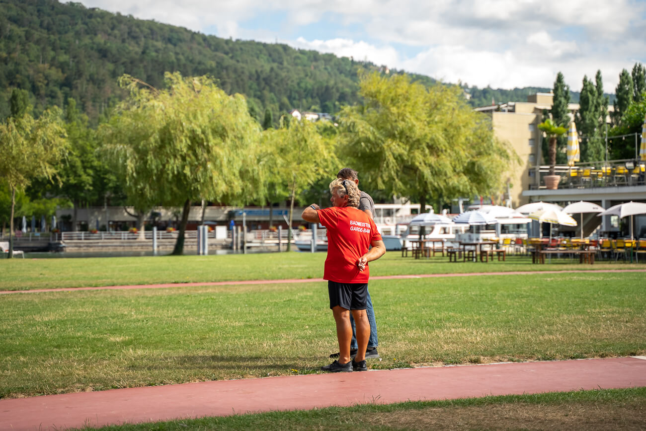 Deux personnes se tiennent ensemble sur un terrain herbeux près d'un chemin. L'une d'elles porte une chemise rouge portant l'inscription