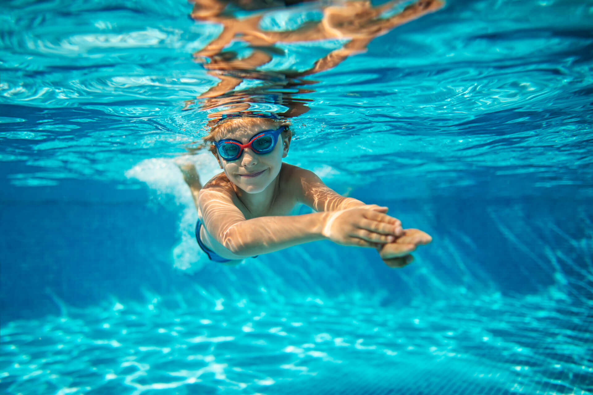 Ein Kind, das eine Schwimmbrille trägt, schwimmt in einem klaren, blauen Schwimmbecken unter Wasser und streckt die Arme nach vorne aus.