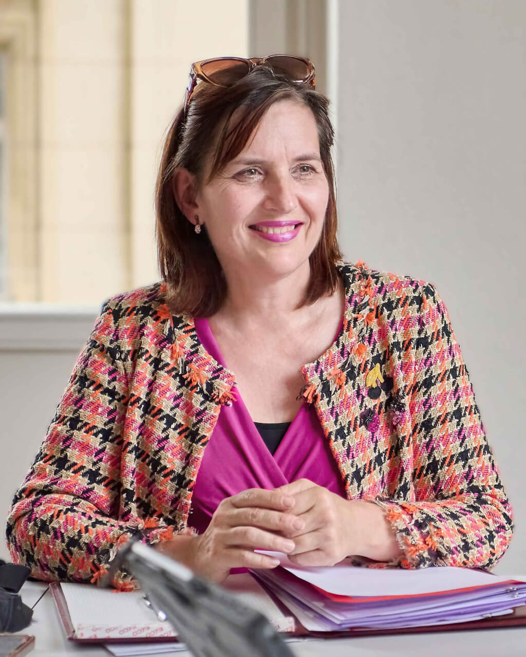 Eine Frau mit schulterlangen braunen Haaren, die eine bunte Jacke und ein rosa Oberteil trägt, sitzt an einem Schreibtisch mit Papieren und einem Notizbuch und lächelt.