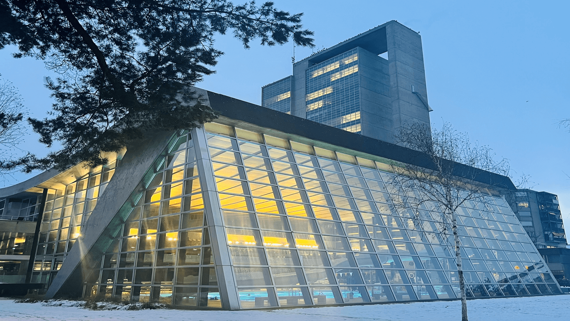Un bâtiment moderne avec une grande façade en verre est éclairé de l'intérieur au crépuscule, avec une architecture angulaire et une haute tour en béton à l'arrière-plan, entourée de neige légère.