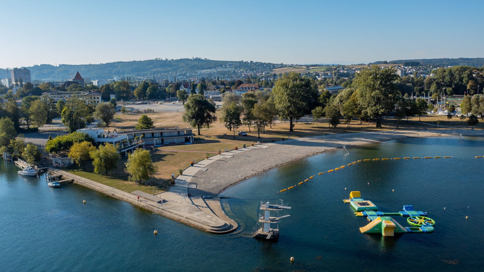 Vue aérienne d'un parc au bord d'un lac avec une plage de sable, des arbres, des bâtiments, une aire de jeux flottante et un paysage urbain lointain sous un ciel bleu clair.