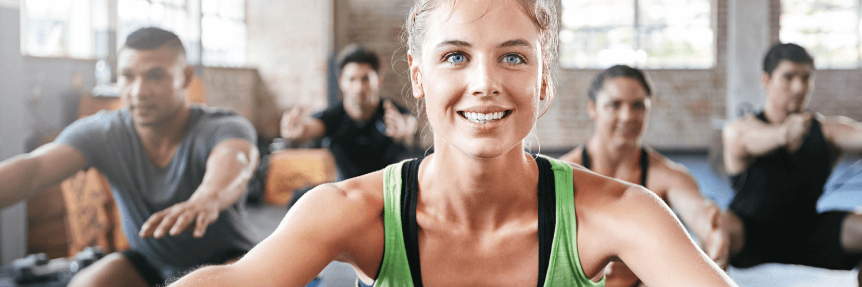 Eine Gruppe von Menschen, die in einer Halle trainieren, mit einer Frau in einem grünen Tank-Top, die vorne lächelt, und anderen, die sich hinter ihr strecken.