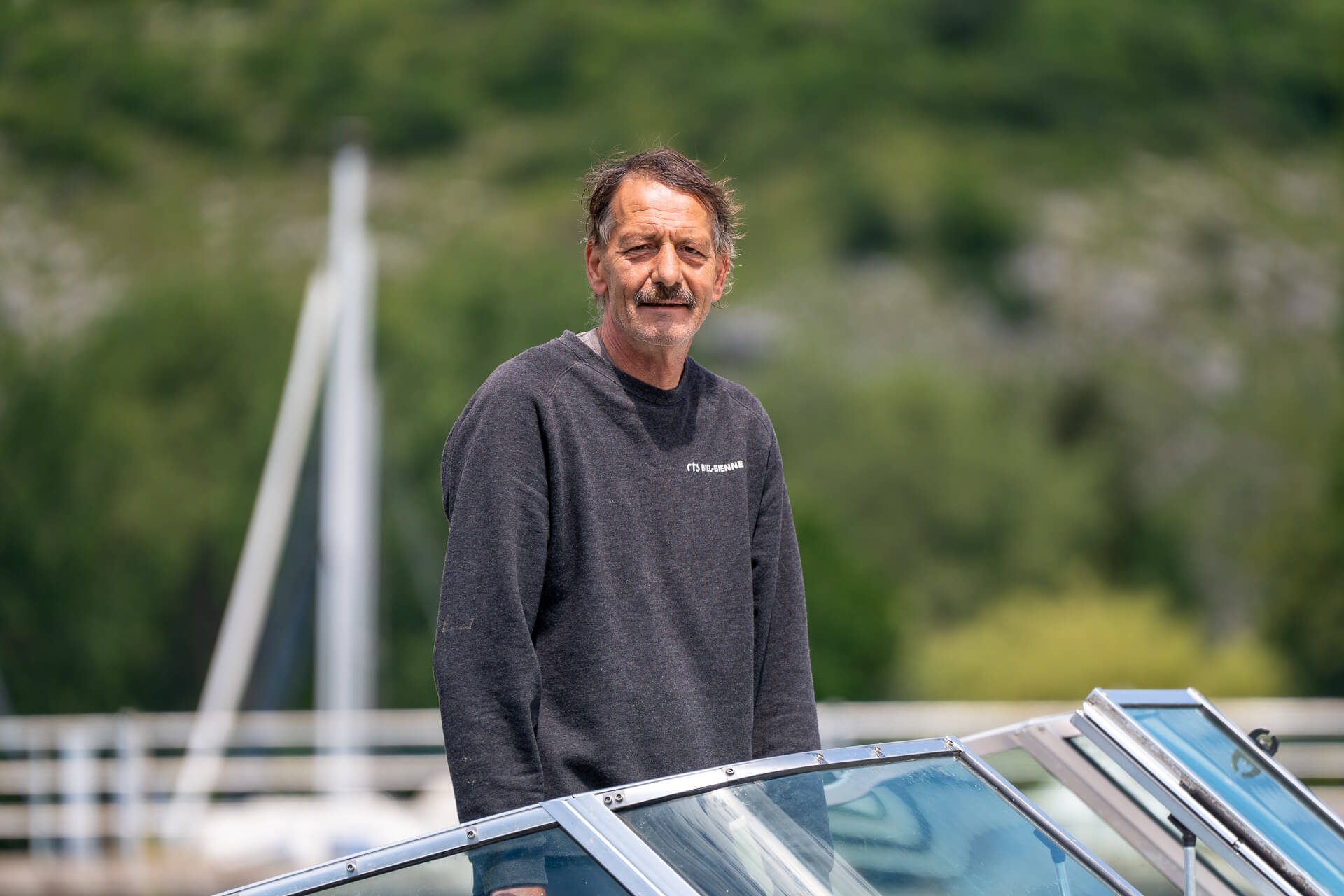 Ein Mann mittleren Alters mit Schnurrbart und dunklem Sweatshirt steht auf einem Boot im Freien, hinter ihm Grünpflanzen und verschwommene Segelbootmasten, die einen Moment aus dem Alltag vom Leiter Eismeister in der Tissot Arena reflektieren.