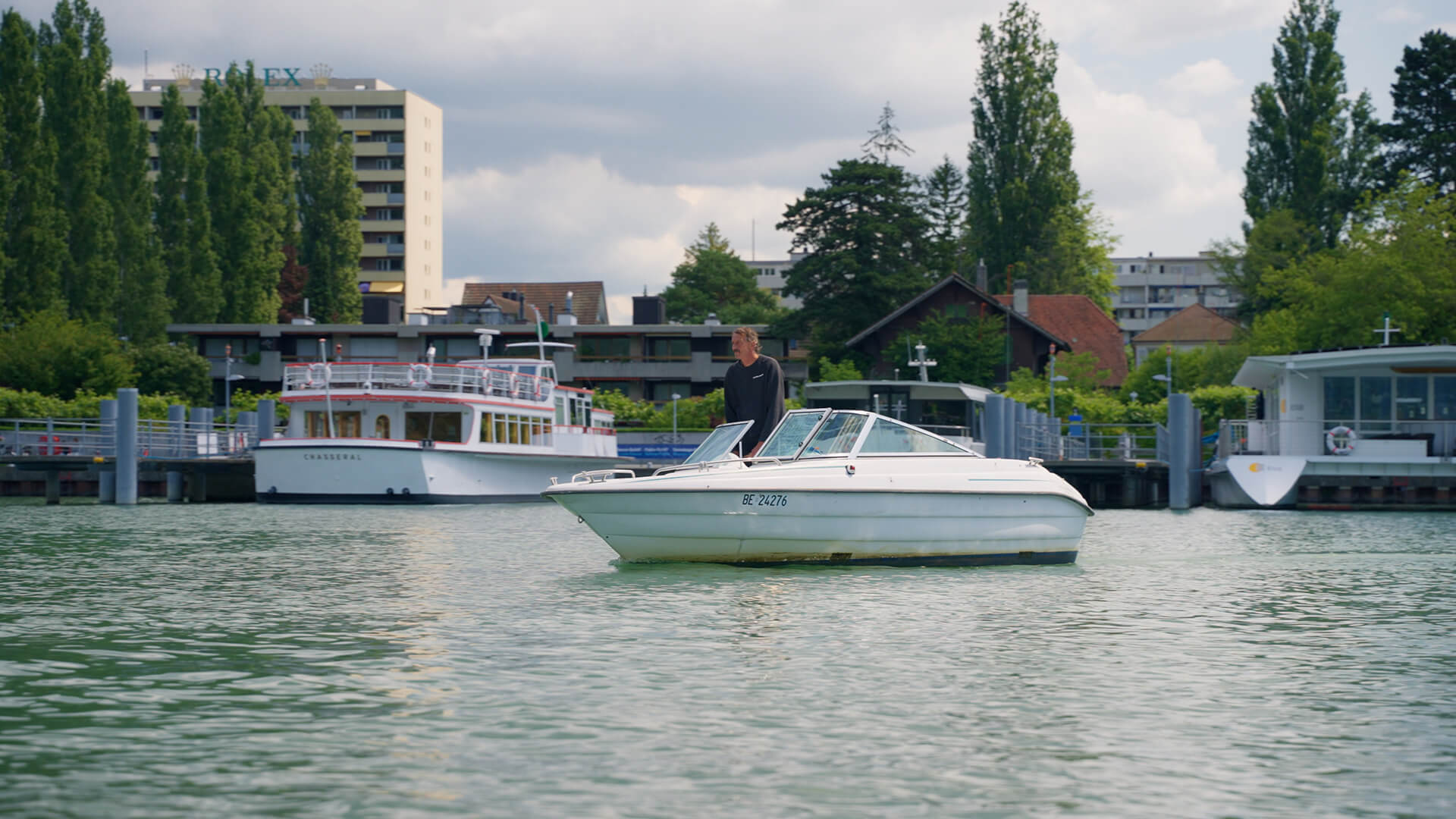 Une personne se tient sur un petit bateau à moteur blanc dans un plan d'eau calme, avec des bateaux plus grands, des arbres et des bâtiments en arrière-plan - rappelant le quotidien du responsable maître glace à la Tissot Arena.