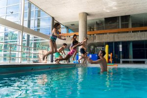 Des enfants en maillot de bain sautent dans une piscine couverte où un adulte est prêt à les rattraper ; des structures de jeu et des fenêtres en verre sont visibles à l'arrière-plan.