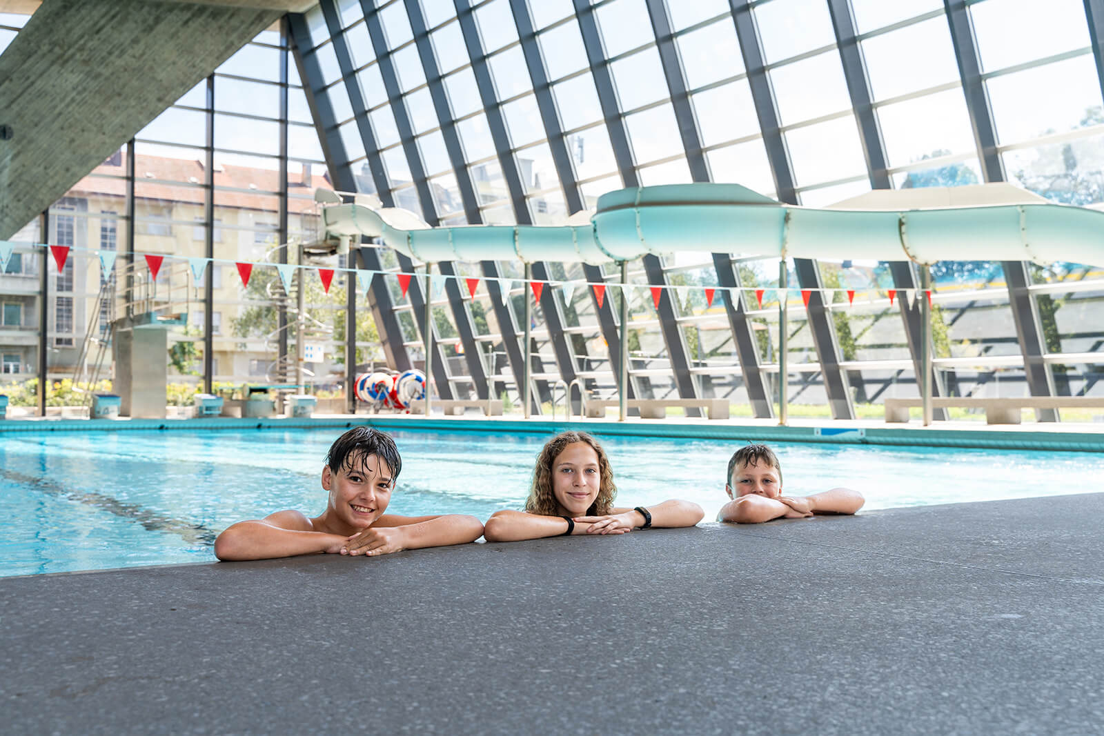 Trois enfants se reposent au bord d'une piscine couverte avec un grand toboggan aquatique et des baies vitrées en arrière-plan.