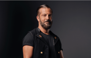 Ein Mann mit zurückgekämmtem Haar und gestutztem Bart steht vor einem dunklen Hintergrund und trägt eine ärmellose schwarze Weste über einem schwarzen T-Shirt.