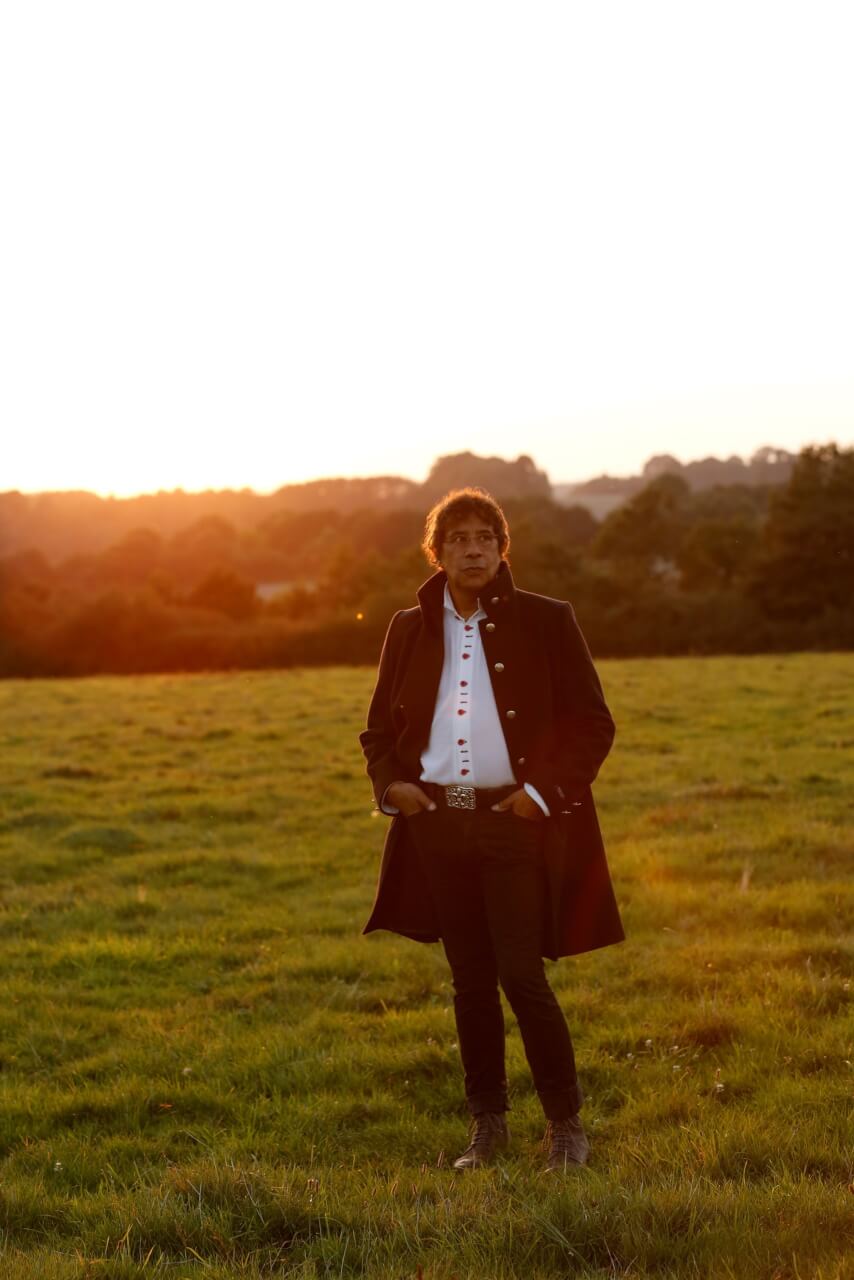Eine Person in einem langen dunklen Mantel steht auf einer Wiese bei Sonnenuntergang, mit Bäumen und Hügeln im Hintergrund.