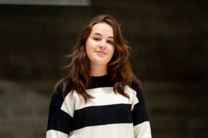 Une femme aux longs cheveux bruns portant un pull rayé noir et blanc se tient devant un fond uni et sombre, capturant l'esprit composé du quotidien du responsable maître glace à la Tissot Arena.