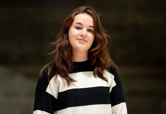 Une femme aux longs cheveux bruns portant un pull rayé noir et blanc se tient devant un fond uni et sombre, capturant l'esprit composé du quotidien du responsable maître glace à la Tissot Arena.