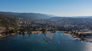 Vue aérienne d'une ville côtière avec un port de plaisance, entourée de collines boisées et de montagnes sous un ciel bleu clair.