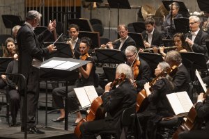 Ein Dirigent leitet ein Orchester mit Musikern, die auf einer Konzertbühne Streich- und Blasinstrumente spielen.