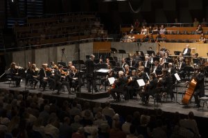 Ein Sinfonieorchester spielt unter der Leitung eines Dirigenten auf der Bühne, wobei die Musiker verschiedene Instrumente spielen und das Publikum davor sitzt.