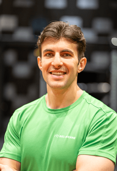 Un homme aux cheveux bruns courts, vêtu d'une chemise de sport vert vif, se tient à l'intérieur, souriant à l'appareil photo, les bras croisés.