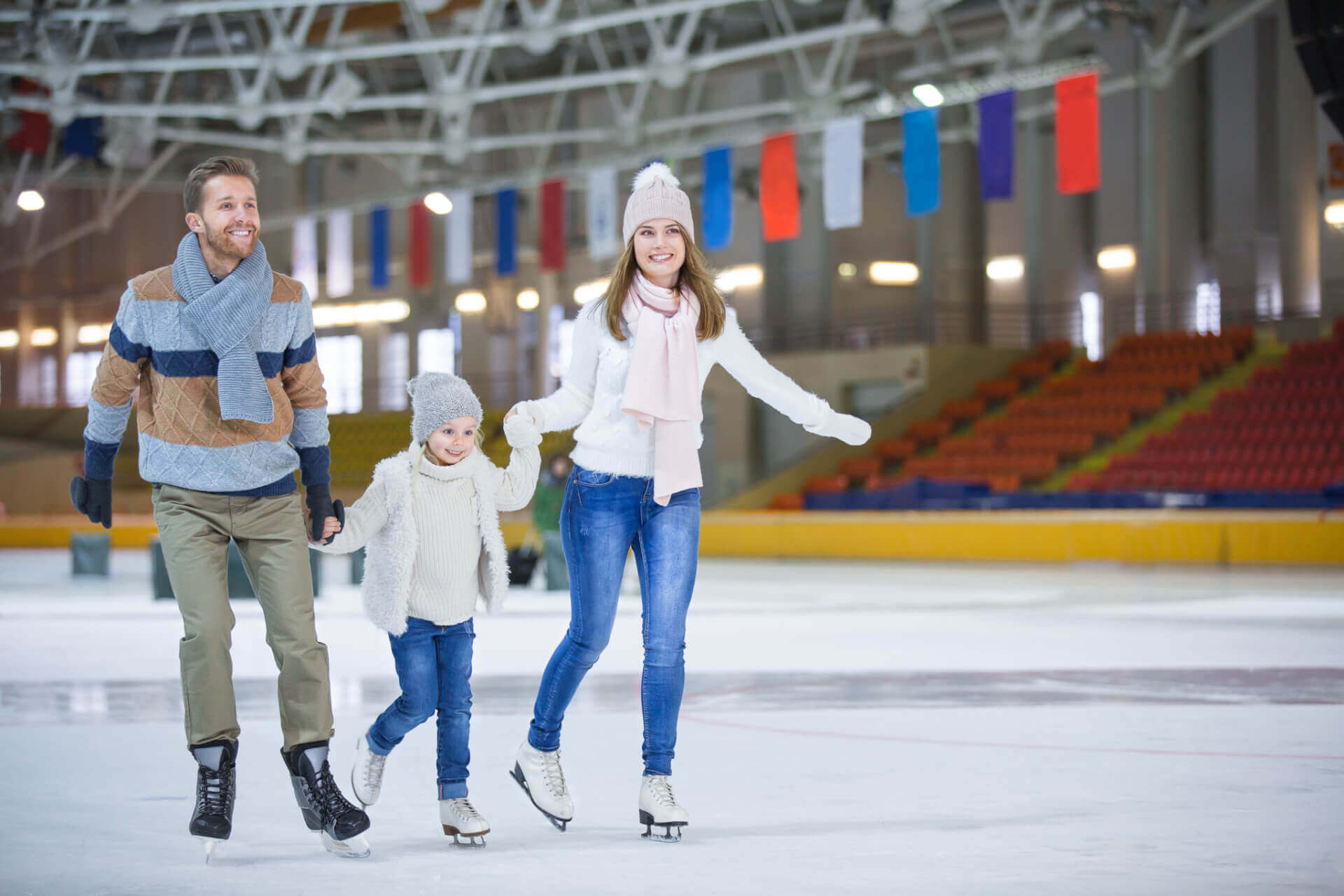 Un homme, une femme et un enfant patinent ensemble à l'intérieur en se tenant par la main, avec des sièges de stade vides et des drapeaux en arrière-plan.