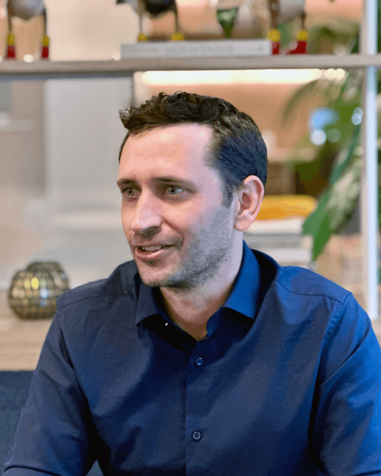 Ein Mann mit kurzen dunklen Haaren und einem Bart, der ein marineblaues Hemd trägt, sitzt in einem Raum mit Regalen und Dekorationsgegenständen im Hintergrund.