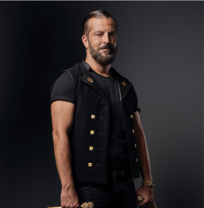 Un homme barbu aux cheveux gominés se tient sur un fond sombre, vêtu d'un t-shirt noir, d'une veste noire sans manches avec des boutons dorés, et tenant un objet dans sa main droite.
