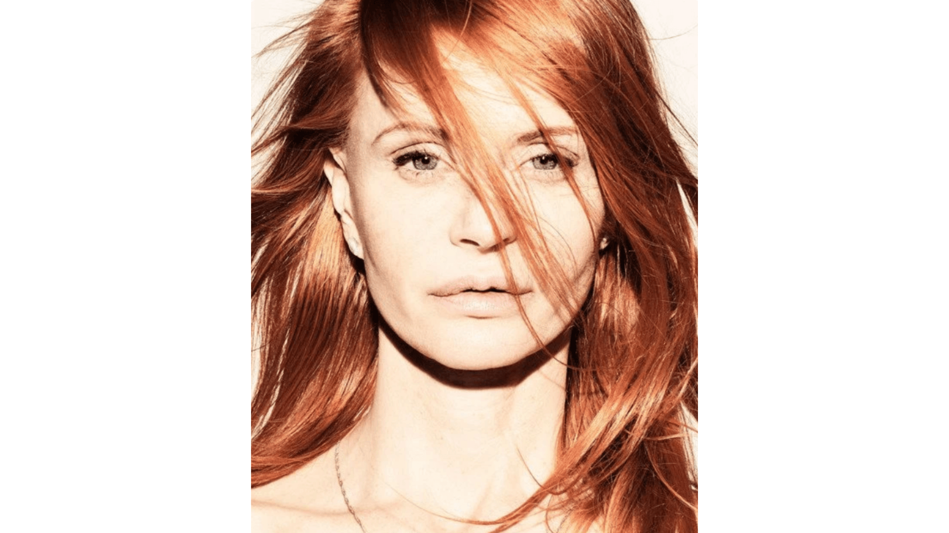 Eine Frau mit langen roten Haaren blickt in die Kamera, wobei ihr einige Haarsträhnen ins Gesicht fallen. Der Hintergrund ist weiß und die Beleuchtung ist hell.