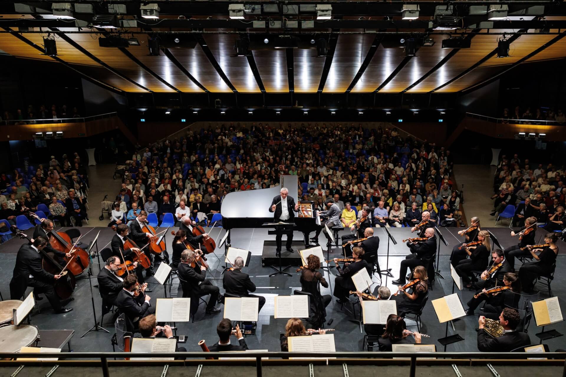 Un orchestre symphonique se produit sur scène devant un public assis dans une salle de concert, sous la direction d'un chef d'orchestre.