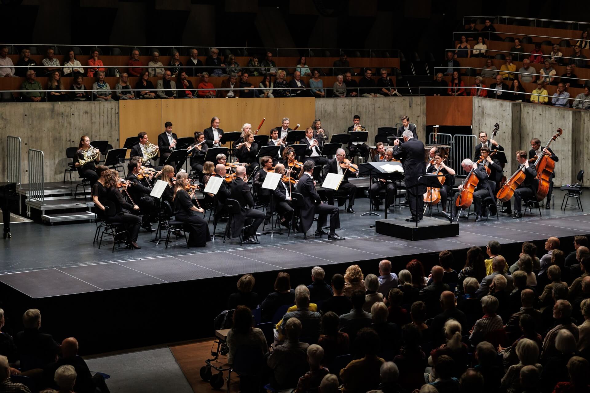 Ein Sinfonieorchester spielt auf der Bühne eines Konzertsaals, mit einem Dirigenten am Dirigentenpult und einem Publikum, das vor dem Orchester sitzt.