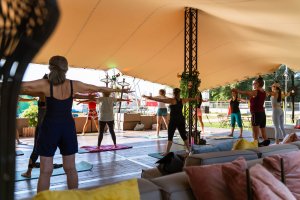 Un groupe de personnes participe à un cours de yoga ou de fitness sous un grand auvent en plein air, debout sur des tapis, les bras tendus.