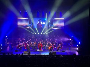Une chanteuse vêtue d'un manteau rouge se produit sur scène avec un orchestre et un chef d'orchestre, éclairés par des lumières de scène, avec des écrans géants affichant la chanteuse derrière eux.