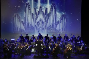 Un orchestre symphonique se produit sur scène avec un chœur derrière lui ; un paysage urbain futuriste est projeté en arrière-plan dans des couleurs vives.
