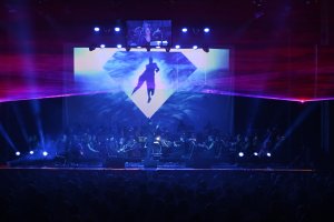 Un orchestre se produit sur scène avec des éclairages bleus et violets ; un grand écran derrière eux affiche la silhouette d'une personne à l'intérieur d'un losange.