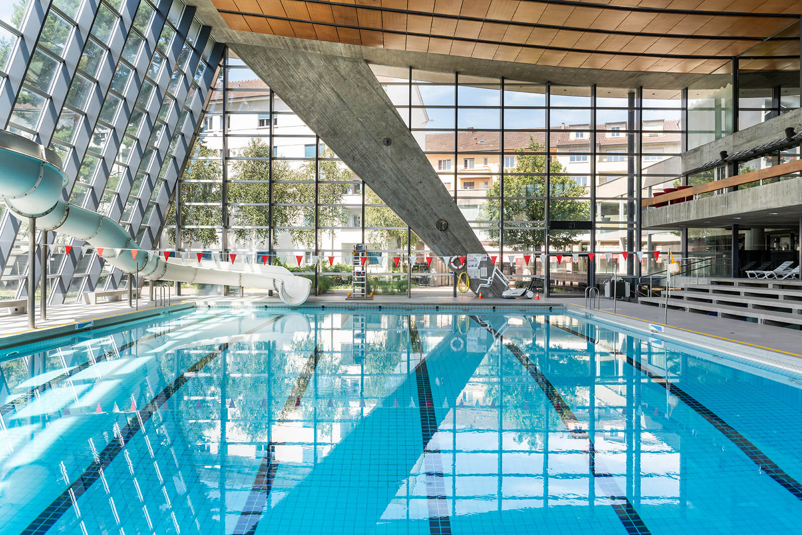 Hallenbad mit klarem blauen Wasser, grossen Fenstern, Wasserrutschen auf der linken Seite und Liegestühlen auf der rechten Seite. Gebäude und Bäume außerhalb sichtbar.