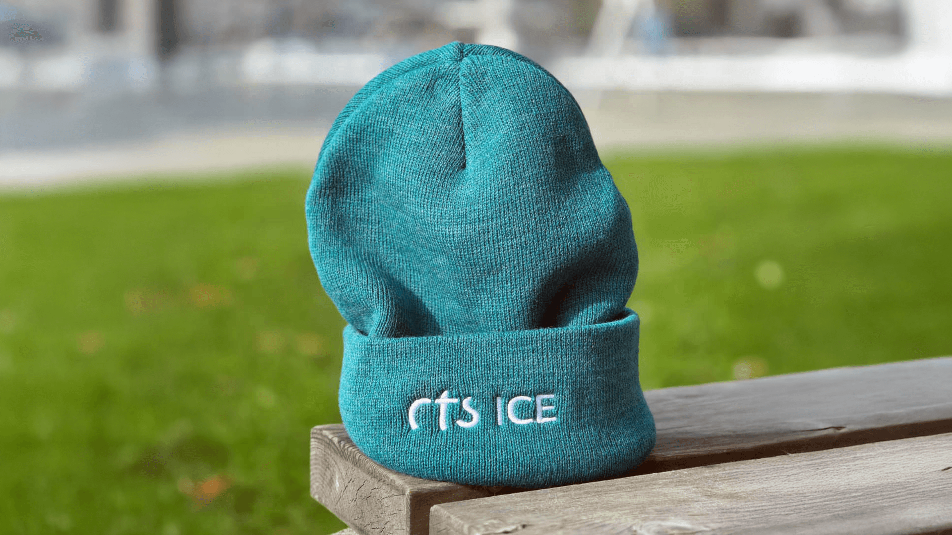 Un bonnet sarcelle avec les mots ITS ICE brodés en lettres blanches est posé sur un banc en bois avec de l'herbe et des pavés en arrière-plan.