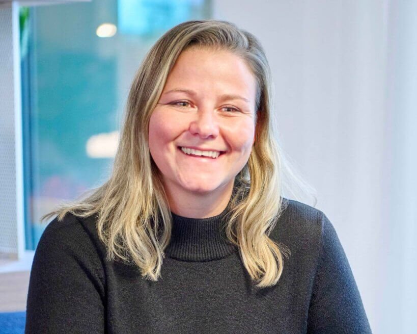 Eine Person mit langen blonden Haaren und einem schwarzen Pullover sitzt an einem Tisch und lächelt mit gefalteten Händen.