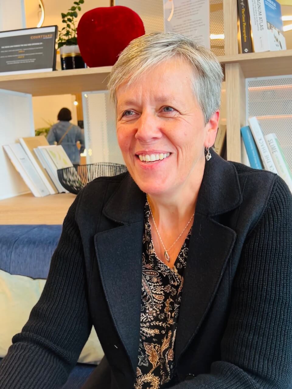 Eine Frau mit kurzen grauen Haaren und einem schwarzen Blazer sitzt lächelnd in einem Haus mit Bücherregalen und Dekoration im Hintergrund.