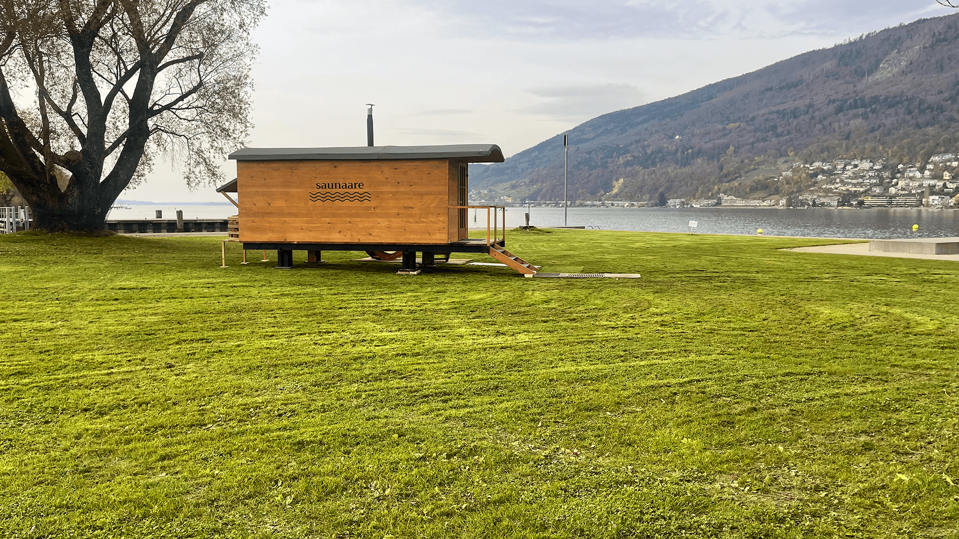 Une petite cabine de sauna en bois sur roues est garée sur un terrain herbeux près d'un lac, avec des collines et des maisons visibles à l'arrière-plan.