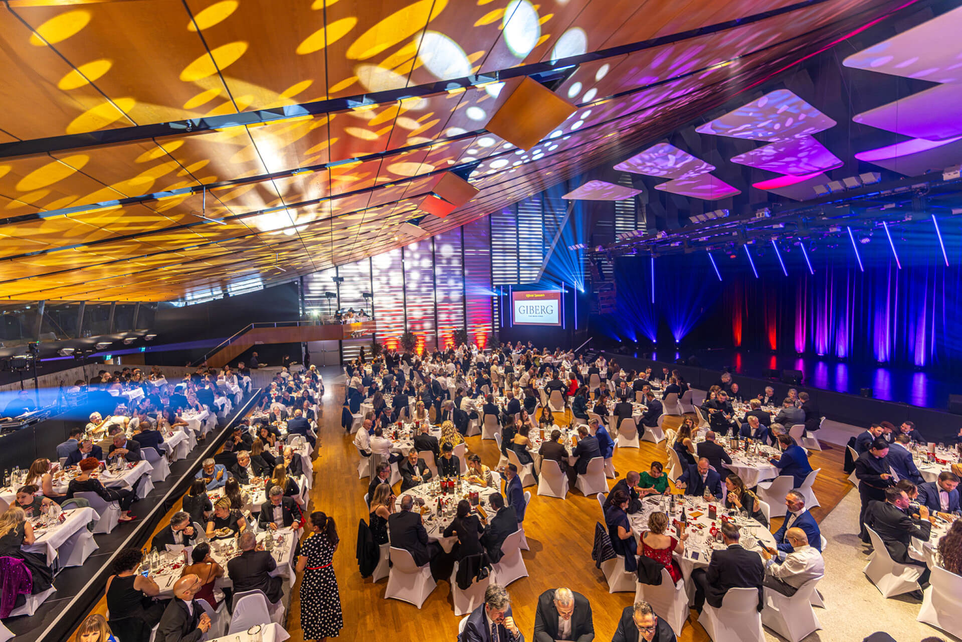 Grand banquet formel avec de nombreux invités assis dans un lieu spacieux et moderne avec un éclairage dramatique et une scène affichant un écran sur lequel on peut lire GEBA Gala.